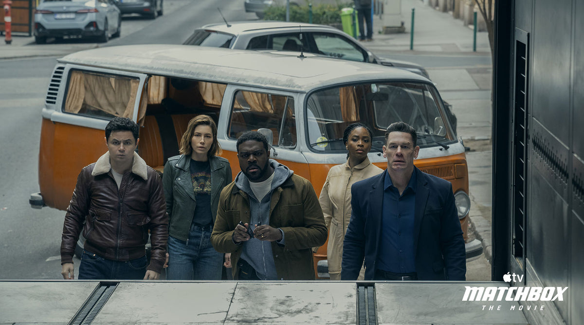 Translation missing: es.Five actors walk forward in front of a vintage orange and white van on a city street, with cars and a sidewalk visible in the background. Matchbox The Movie Apple TV logo appears in the bottom right corner