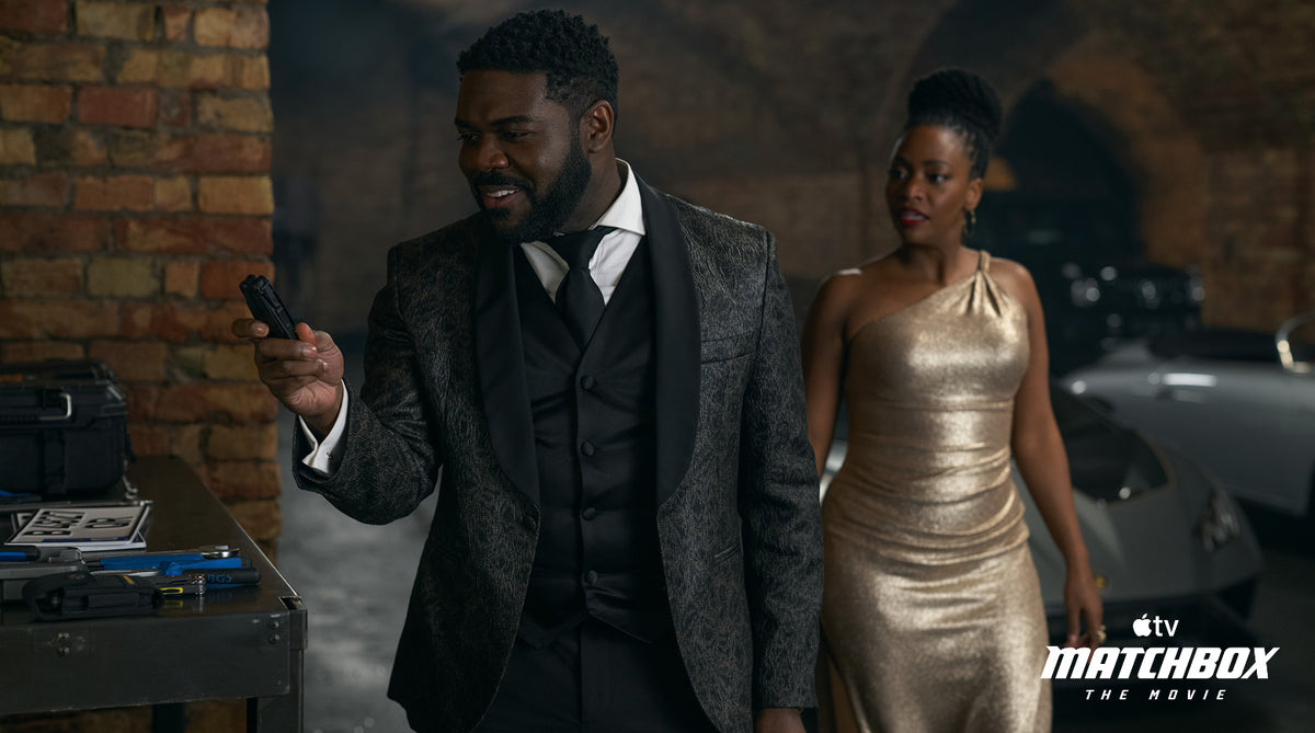 Translation missing: es.A scene from Matchbox The Movie. Crosby, in a black patterned tuxedo, smiles as he holds up a black Leatherman Surge multi-tool. A woman in a gold one-shoulder gown walks up behind him. The background has a brick wall, a workbench with tools on the left, and parked sports cars in a dim garage. An Apple TV Matchbox The Movie watermark is in the bottom right corner