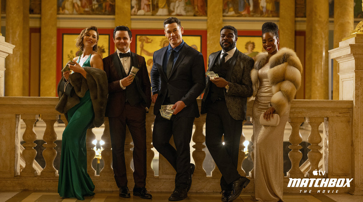Translation missing: es.Five cast members of Matchbox The Movie stand together on an ornate balcony in a grand, classical building. They are dressed in formal evening wear, with one in a green gown, one in a brown suit, one in a dark suit, one in a brown patterned suit, and one in a gold sparkly gown with a fur coat