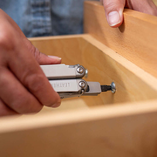 Translation missing: es.Hand using a Stainless Steel Leatherman Wave® Plus multitool on a wooden drawer, showcasing its practical features