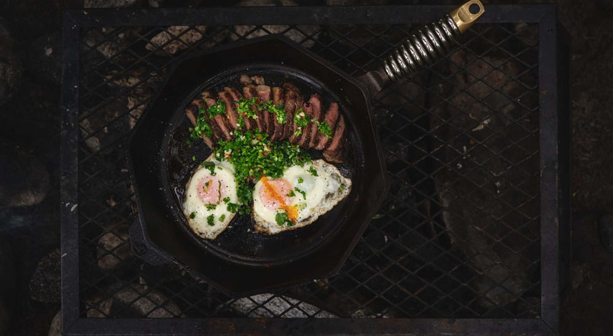 A cast iron skillet on a grill, featuring sunny-side up eggs, sliced meat, and fresh herbs.