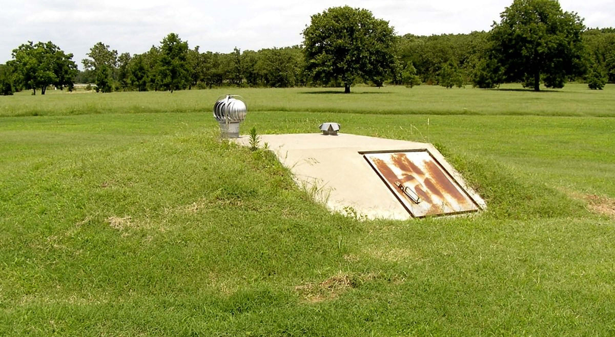 A concrete access hatch set in a grassy area, surrounded by trees, likely leading to an underground bunker.