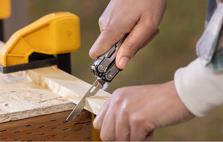 Translation missing: en.Person using the saw on a Black DLC and Stainless Steel Leatherman multitool to cut wood, demonstrating practical skills in a DIY woodworking project