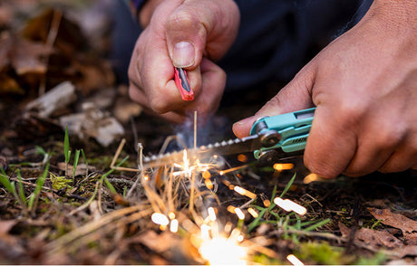 Translation missing: en.Hands using the ferro rod on a Aqua Stainless Leatherman Signal multi-tool to create sparks from dry grass in a forest setting, ideal for outdoor survival skills