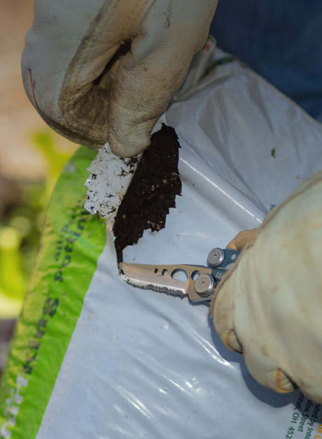 Translation missing: es.Person using a Denim Skeletool® to open a bag of soil, showcasing the utility of this versatile tool in gardening tasks