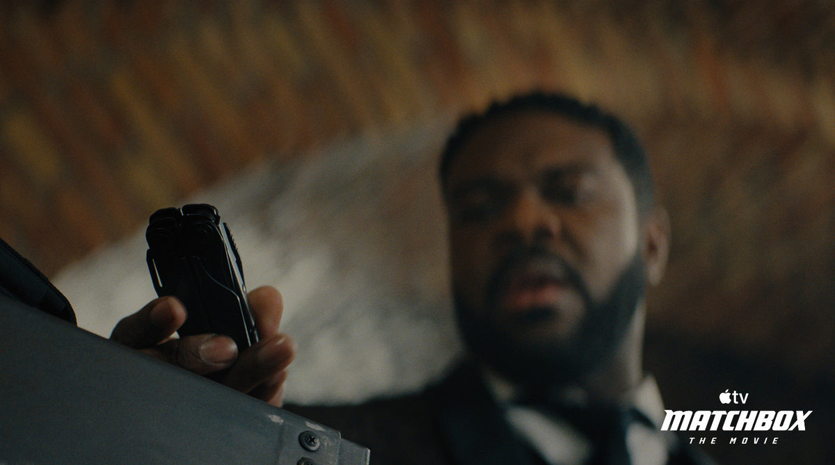 Translation missing: es.A close-up shot of the character Crosby in a black suit holding a black Leatherman Surge, with a blurred brick wall background from Matchbox The Movie. Apple TV Matchbox The Movie watermark appears in the bottom right corner