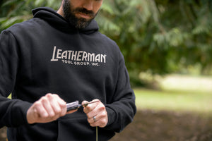 Translation missing: en.A person wearing a black Leatherman Blade &amp; Grain hoodie using a Leatherman multitool to cut a piece of rope outdoors, showing the front logo of the hoodie that reads “Leatherman Tool Group, Inc.”