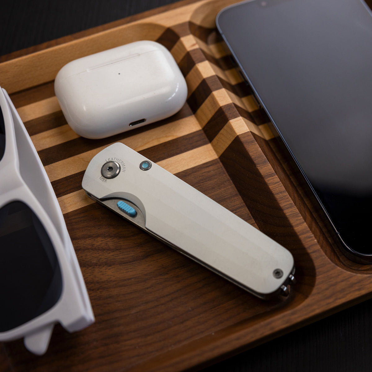Translation missing: en.Leatherman Blazer Alpine folding knife in closed position placed in a wooden valet tray alongside wireless earbuds, a smartphone, and sunglasses, highlighting everyday carry essentials