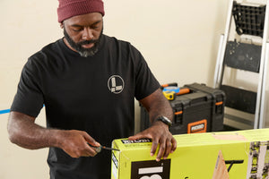 Translation missing: en.A person wearing a black Leatherman Classic Tee using the blade on a Wave Alpha in Cascadia color to open a large box in a workshop while standing near a ladder and tool case