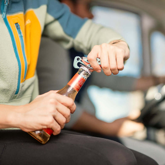 Translation missing: es.Person using a Paradise Skeletool® CX to open a bottle in a car, showcasing its multi-tool functionality
