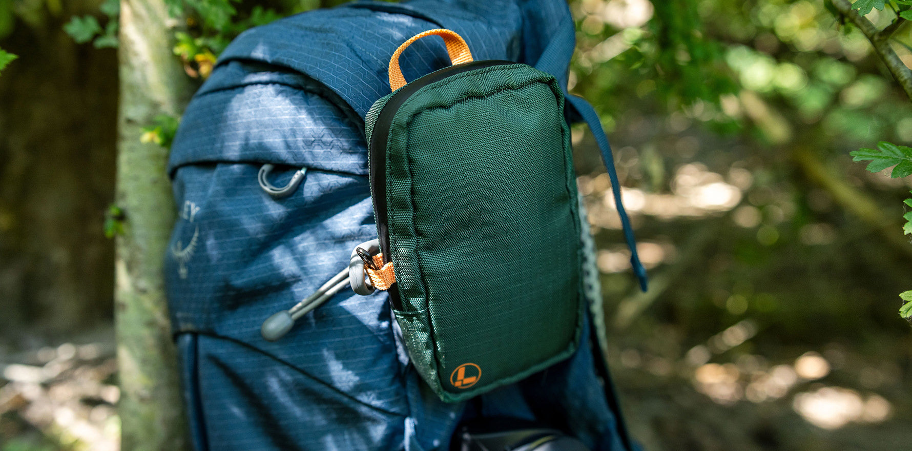 A Leatherman Tool Pouch in Hunter color is clipped to the shoulder strap of a blue hiking backpack, resting in an outdoor forest setting with sunlight filtering through the trees.