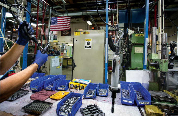 Translation missing: en.Worker assembling parts in a manufacturing facility with tools and organized bins, American flag in the background