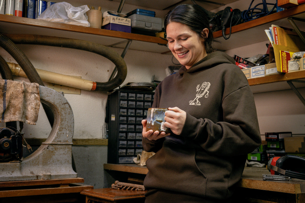 Translation missing: en.A person wearing a brown Leatherman Mr. Wave hoodie standing in a workshop holding a ceramic mug, smiling while surrounded by tools and equipment
