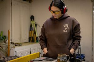 Translation missing: en.A person wearing a brown Leatherman Mr. Wave hoodie standing in a workshop with safety glasses and ear protection, looking down at a workbench with tools and a ceramic mug