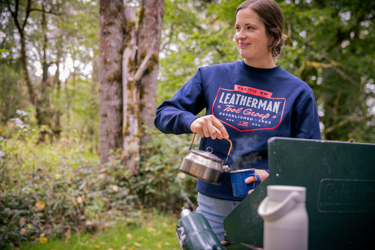 Translation missing: en.A person wearing a navy blue Leatherman PNW Crewneck sweatshirt pouring hot water from a kettle into a mug at a campsite surrounded by trees