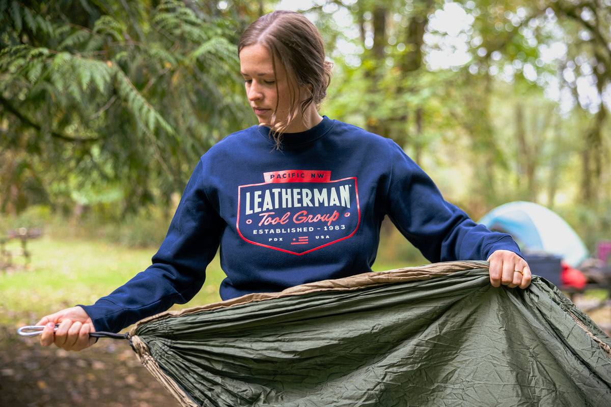 Translation missing: en.A person wearing a navy blue Leatherman PNW Crewneck sweatshirt setting up a hammock at a forest campsite with a tent in the background