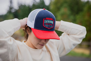 Translation missing: es.A person adjusting a navy Leatherman PNW Trucker Hat with a white mesh back and red brim while outdoors