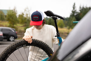 Translation missing: es.A person wearing a navy Leatherman PNW Trucker Hat with a white mesh back and red brim loading a blue mountain bike onto a vehicle