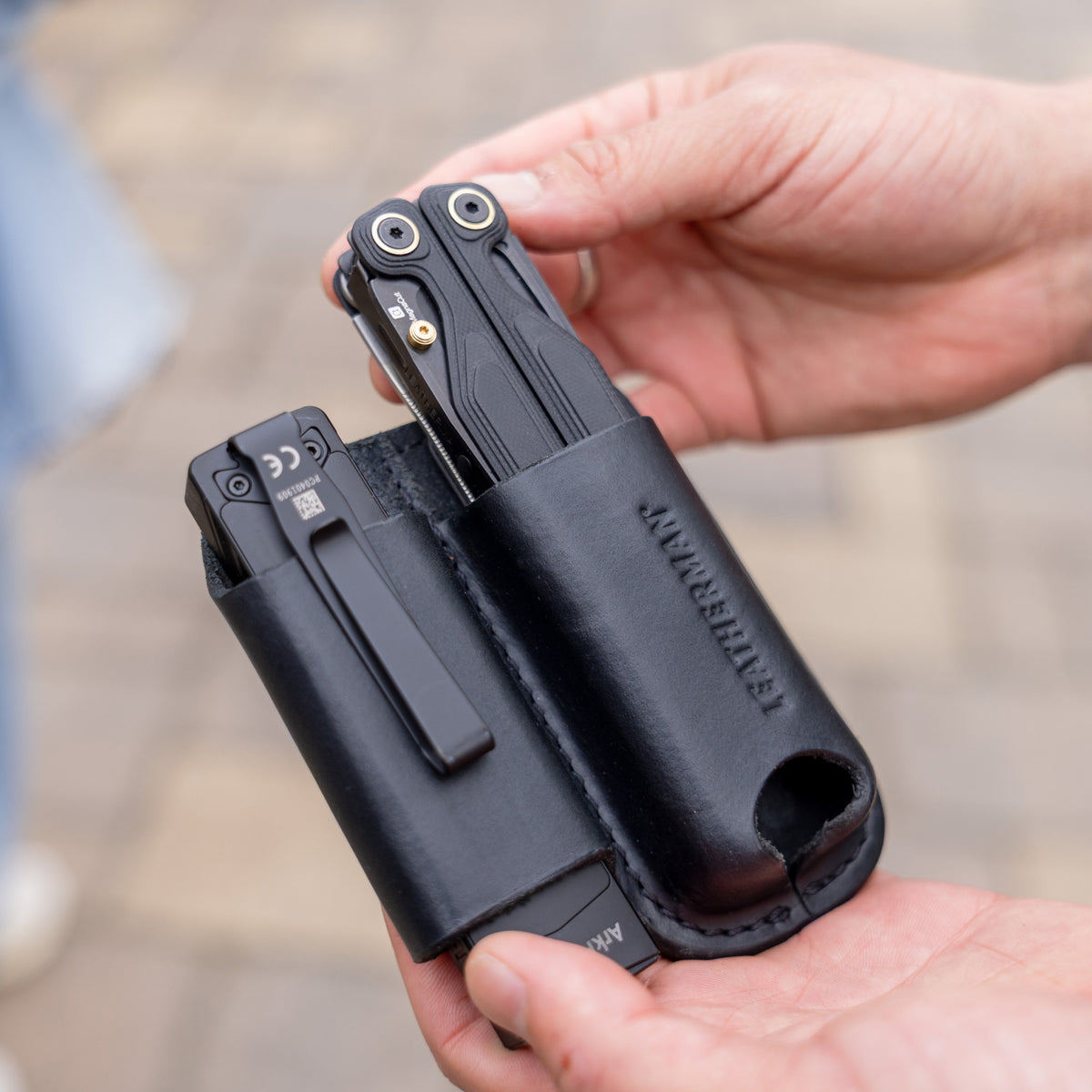 Translation missing: es.Hands holding a black leather sheath carrying two black multitools outdoors