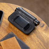 Translation missing: es.Black leather sheath with belt clip holding a small cylindrical flashlight on a wooden surface