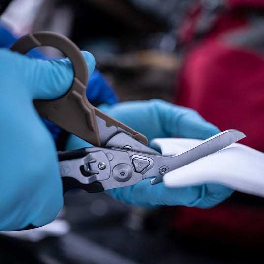 Translation missing: en.Hands in blue gloves using  Coyote Black Raptor® Rescue shears to cut gauze