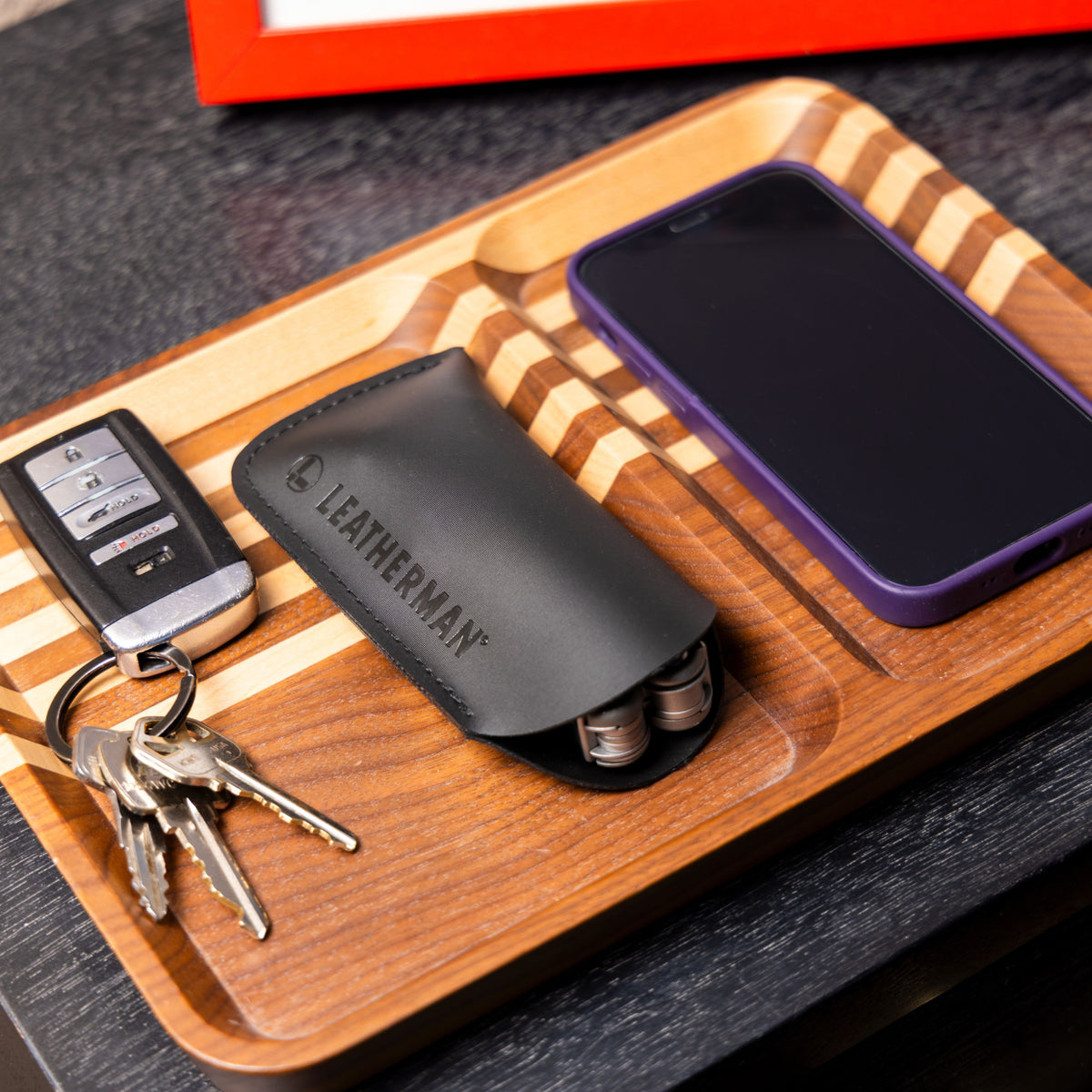 Translation missing: en.A black Leatherman Hypalon tool sleeve holding a multi-tool, neatly placed on a wooden valet tray beside a set of keys and a smartphone with a purple case, showcasing an organized everyday carry setup