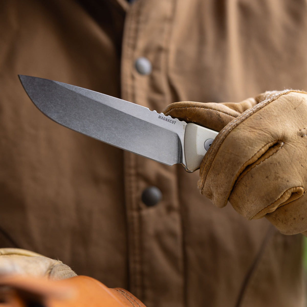 Translation missing: en.Person wearing work gloves and a brown canvas jacket holding a Leatherman Trac Alpine fixed-blade knife outdoors, emphasizing the blade and handle in a close-up view